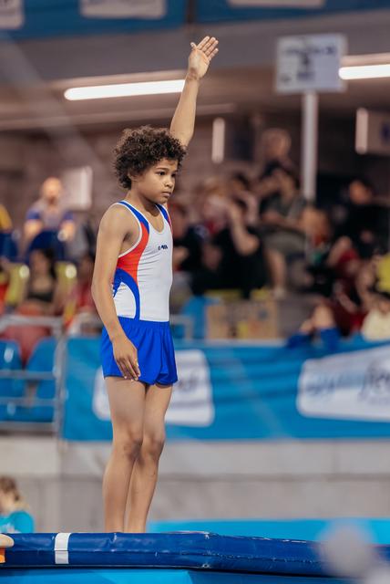 Young gymnast raises hand to signal readiness on trampoline, showing calm focus before their routine in a busy sports hall.