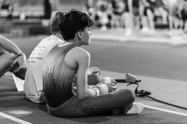 Young gymnast sits cross-legged on the floor, wrists wrapped, watching intently with quiet focus during a gymnastics event.