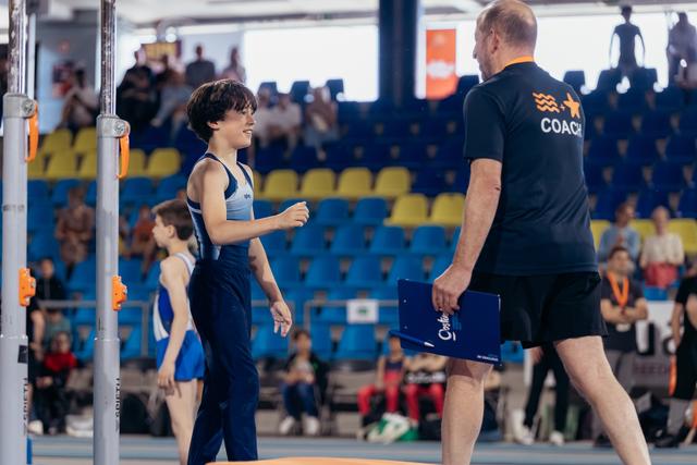 Young male gymnast smiles warmly at his coach holding a clipboard, in a competition arena with blue and yellow seats.