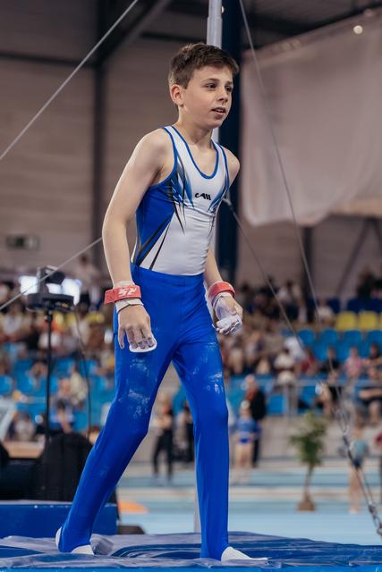 Young male gymnast stands poised on mat, chalk-covered hands, looking aside with calm focus during indoor gymnastics event.