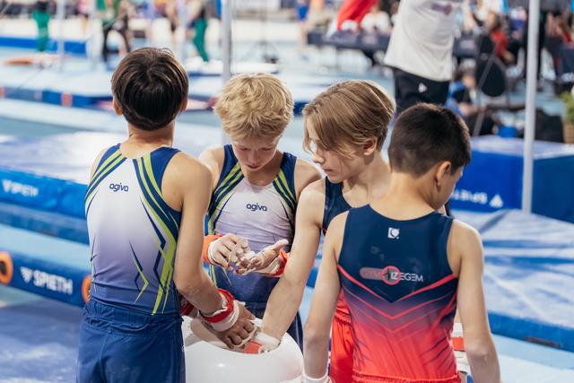 Four young male gymnasts huddle together applying chalk to their hands, focused and preparing before their routine at a gymnastics meet.