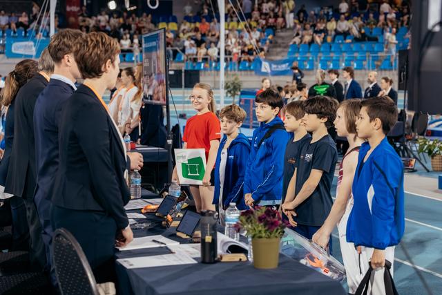 Young gymnasts in blue jackets line up attentively before officials at a registration table inside a packed sports arena.