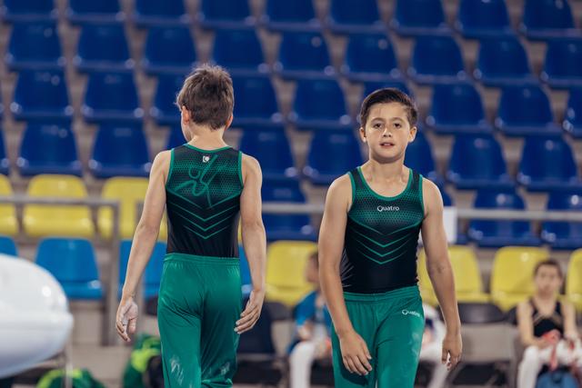 Two young male gymnasts in green Quatro uniforms walk together in an arena, one facing forward with a focused expression.