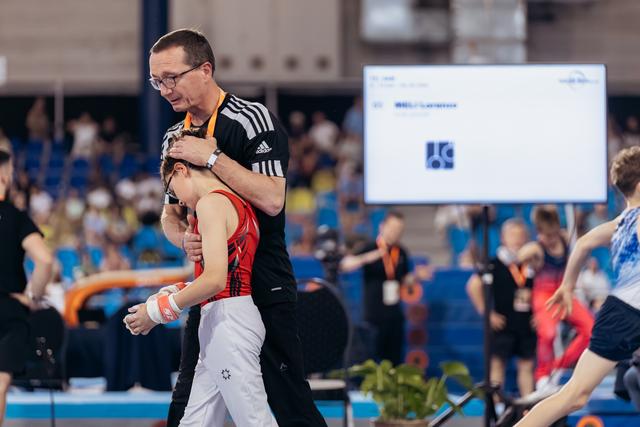 A coach embraces a young gymnast in a red leotard, offering comfort after a performance in a busy arena.
