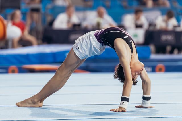 Young gymnast performs a deep backbend on the floor exercise mat during a competition, barefoot with wrist guards.