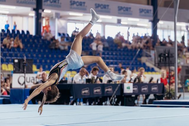 Young gymnast mid-backflip on floor exercise, body fully extended, competing before a packed arena crowd.