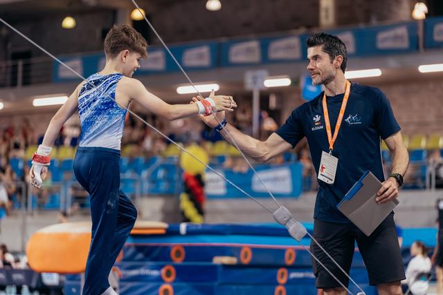 Young male gymnast shakes hands with smiling coach holding clipboard, sharing a proud moment on the competition floor.