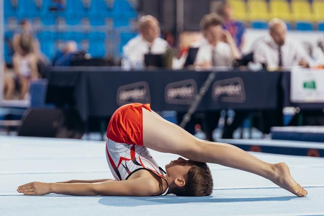 Young male gymnast performs an extreme backbend on the floor mat, judges watching in the background.
