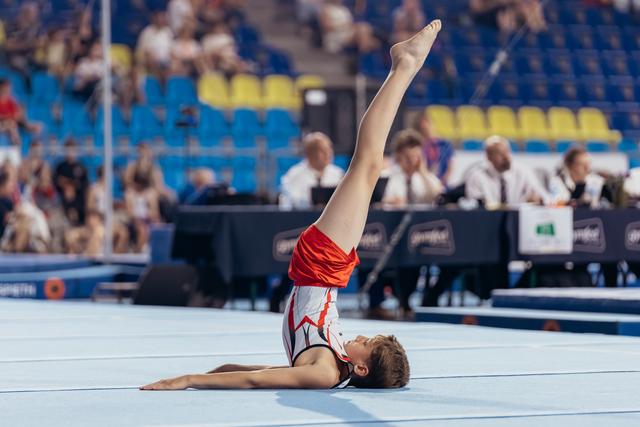 Young male gymnast performs a leg raise on floor, body arched gracefully, during a competition in a packed arena.