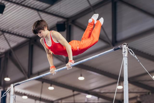 Young gymnast in red uniform grips the horizontal bar with intense focus, legs raised in a pike position indoors.