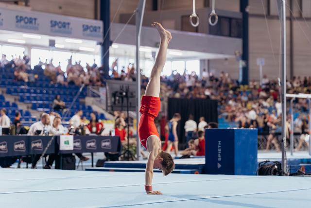 Male gymnast in red leotard holds a perfect handstand on the floor exercise mat before a packed arena crowd.