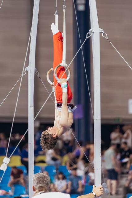 Young male gymnast performs inverted hang on still rings, shirtless in red pants, intense concentration in packed arena.