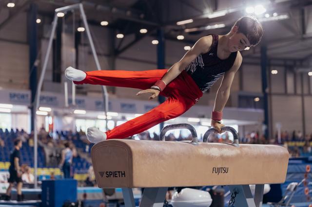 Young male gymnast performs scissor move on pommel horse, leaning forward with focused intensity in packed arena.