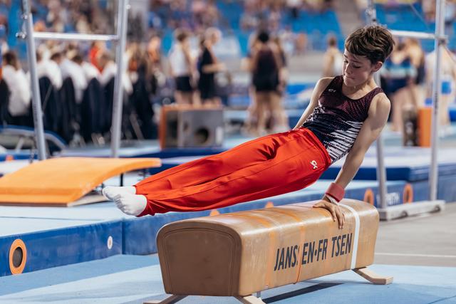 Young male gymnast performs pommel horse routine, body parallel to floor, focused expression, in a competition arena.