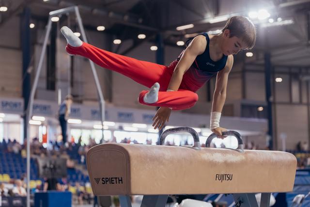 Young male gymnast performs scissor move on pommel horse, legs split wide, intense focus, in a packed competition arena.