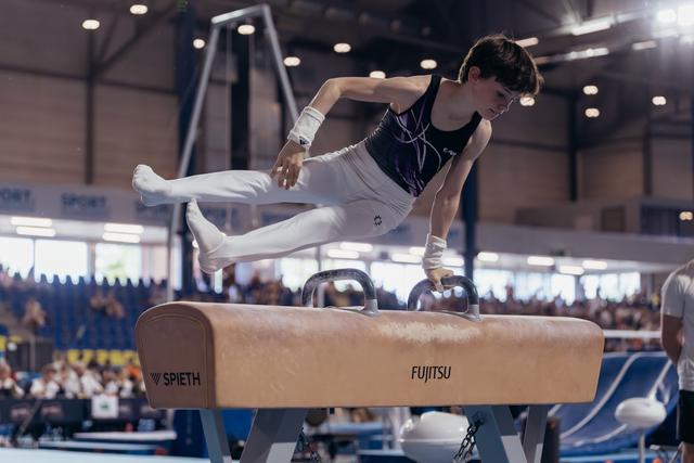 Young male gymnast performs on pommel horse, leaning forward with intense concentration in a packed arena.