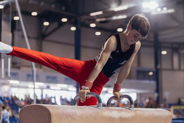 Young male gymnast performs on pommel horse, legs extended sideways, focused expression, in a large indoor gymnastics hall.