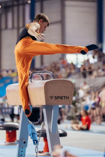 Young male gymnast in orange leotard performs a leg sweep on the pommel horse, focused and controlled.