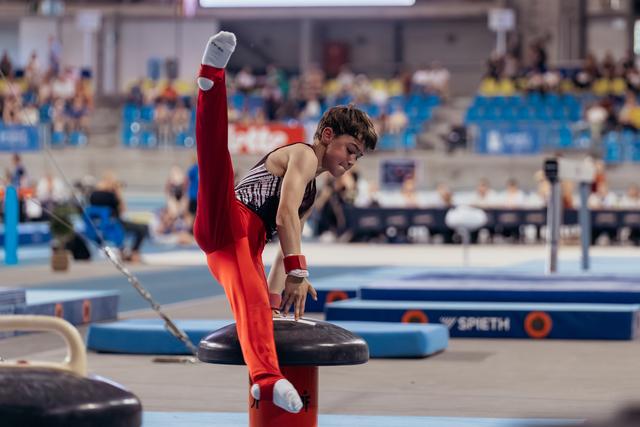 Young male gymnast performs a split on the pommel horse, leaning forward with intense concentration at an indoor competition.