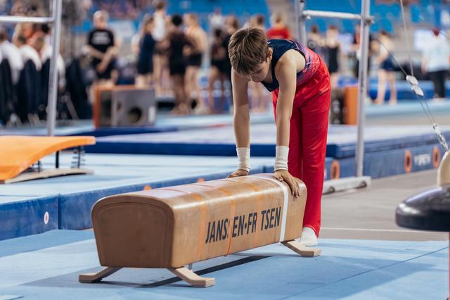 Young male gymnast leans intently over a pommel horse, hands on apparatus, focused before his routine in a busy gym.