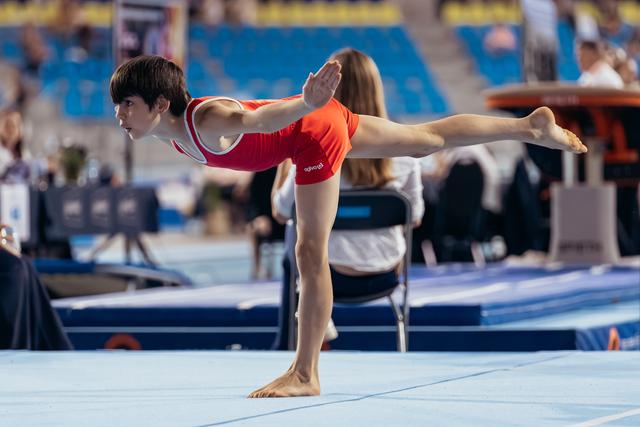Young gymnast in red leotard holds a focused arabesque balance on the floor exercise mat during competition.