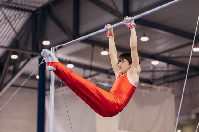 Young gymnast in red leotard hangs from horizontal bar with focused expression, legs extended in a straddle position.