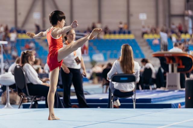 Young gymnast in red leotard performs a high leg raise on the floor exercise mat, focused and precise.