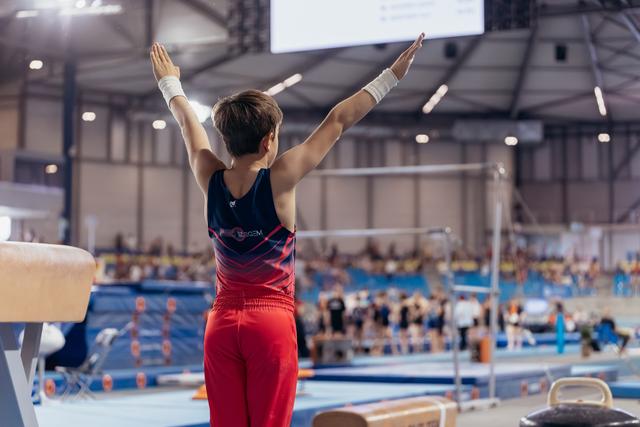 Young male gymnast raises both arms in a triumphant salute inside a large gymnastics arena, wearing red and navy.