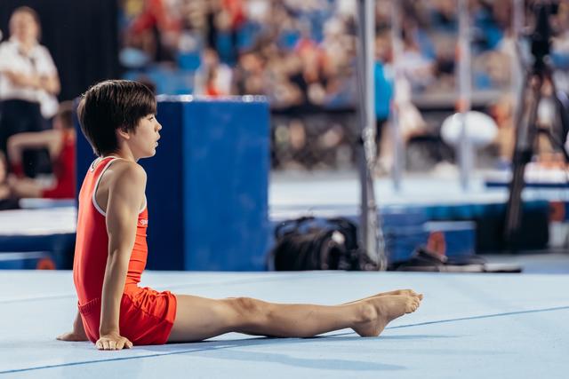 Young gymnast sits in a straddle position on the blue floor mat, composed and focused, during a competition.