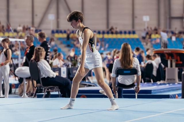 Young gymnast in Belgian colours stands composed on the blue floor, gathering focus before or after a routine.