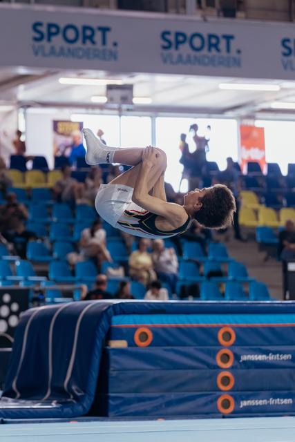 Young male gymnast performs a tight tuck backflip above the trampoline bed, focused and airborne in a packed arena.