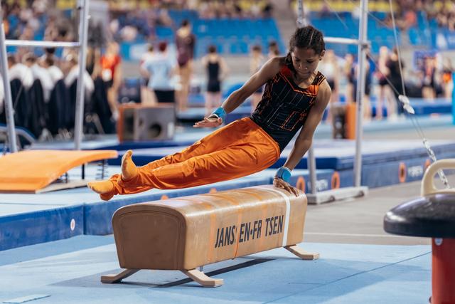Young gymnast in orange uniform vaults over pommel horse with focused intensity at indoor gymnastics competition.
