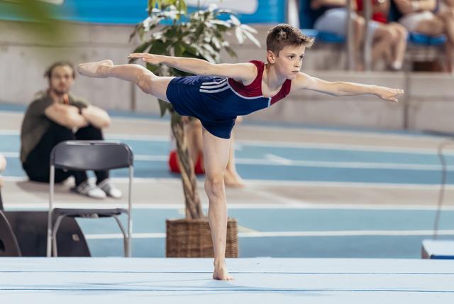 Young male gymnast holds a precise arabesque on floor, arms outstretched, intense focus on his face in an indoor arena.
