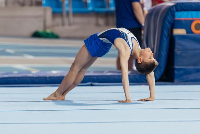 Young gymnast performs a back bridge on the floor exercise mat, arms and legs extended, head tilted back with focus.