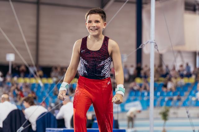 Young male gymnast celebrates with fists clenched and a beaming smile after his routine in a packed arena.