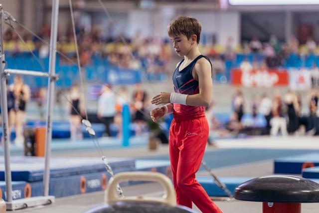 Young male gymnast in red and navy leotard applies chalk to his hands, focused and composed before his routine.