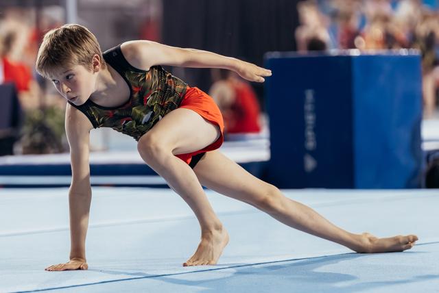 Young gymnast performs a focused floor routine lunge, arms extended, showing intense concentration during competition.