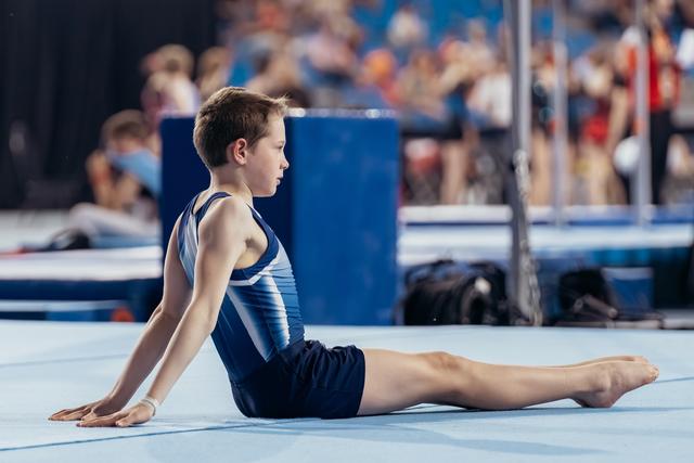 Focused young male gymnast performs a seated stretch on the floor exercise mat during a gymnastics meet.