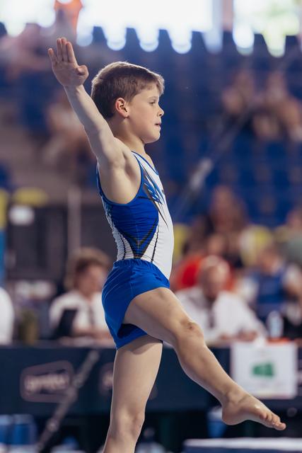 Young boy gymnast mid-leap on floor exercise, arm raised, focused expression, performing at indoor gymnastics meet.