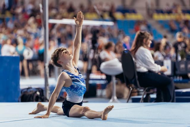 Young gymnast performs a split pose on the floor mat, arm raised gracefully upward, face lifted with focused expression.