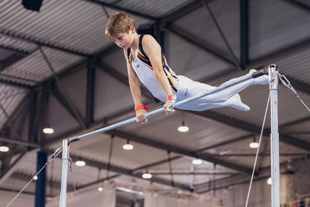 Young male gymnast grips the high bar with intense focus, legs extended mid-routine in an indoor gymnasium.