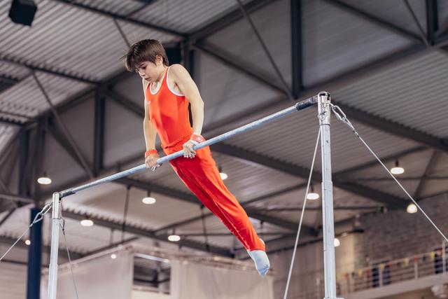 Young male gymnast in red leotard grips the high bar with intense focus, legs extended mid-routine in an indoor gymnasium.