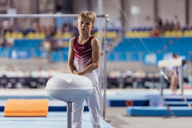 Young male gymnast in red Quatro leotard stands pensively at pommel horse, calm expression, in a large gymnastics arena.