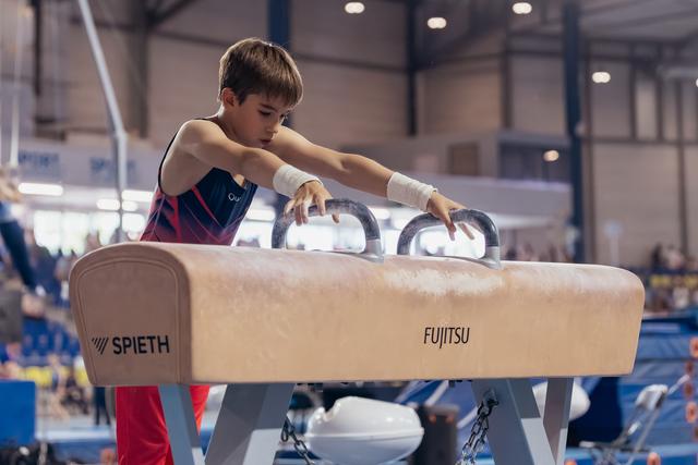 Focused young male gymnast grips pommel horse handles with concentration, wearing wrist guards at an indoor gymnastics meet.