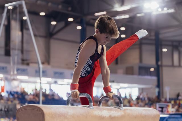 Young male gymnast performs on pommel horse, leg raised high, expression focused and determined, indoor arena setting.