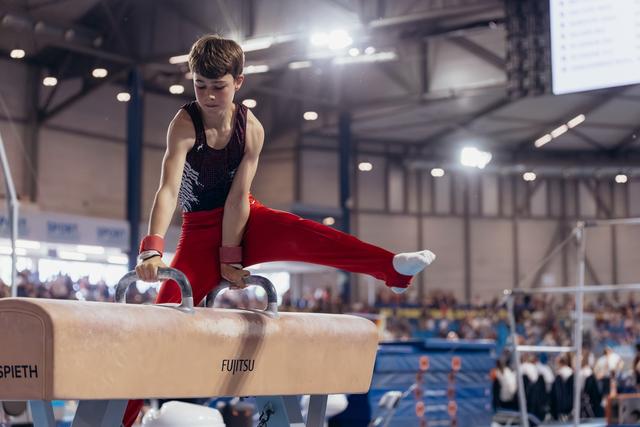 Young male gymnast performs focused pommel horse routine, leg extended, in a large indoor arena with spectators.