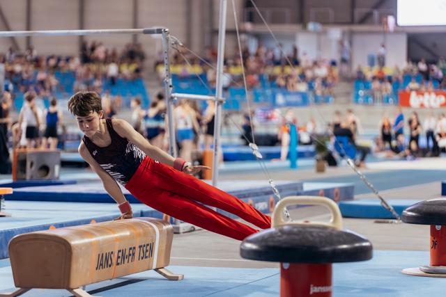 Young male gymnast performs on pommel horse with intense focus, leaning forward in red pants at indoor gymnastics meet.