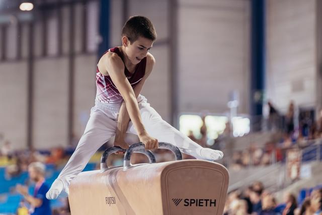 Young male gymnast performs on pommel horse, leaning forward with intense focus during a gymnastics meet.