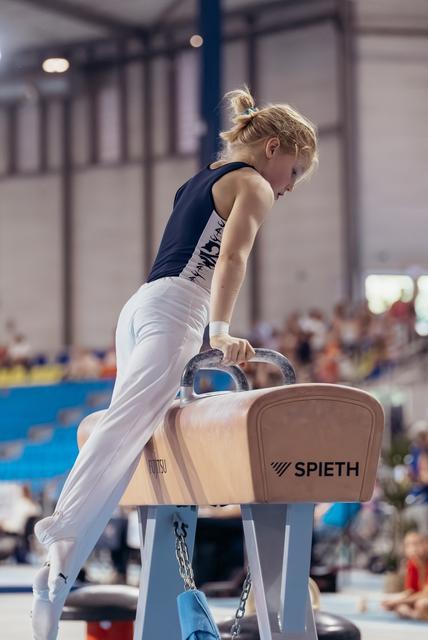 Young male gymnast grips pommel horse handles with intense focus, leaning forward mid-routine at indoor competition.