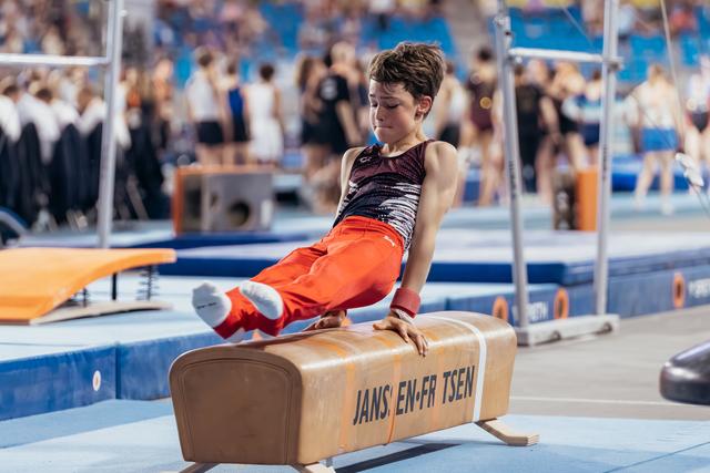 Young male gymnast performs focused pommel horse routine, legs raised, wearing red pants in a busy sports hall.